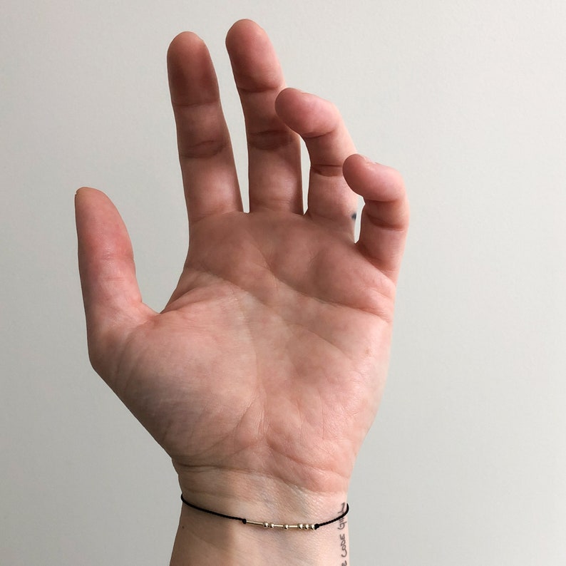 May include: A black string bracelet with gold-coloured beads. The bracelet is worn on a wrist with a small tattoo. The background is a plain, light-coloured wall.