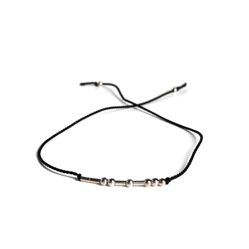 May include: A black cord bracelet featuring silver beads and cylindrical elements. The bracelet is adjustable and set against a white backdrop. The beads are spaced along the cord, creating a minimalist design.
