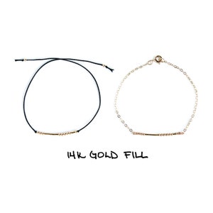 May include: Two bracelets, one with a black cord and one with a gold chain, both with a gold bar and beads. The text "14K GOLD FILL" is written below the bracelets.