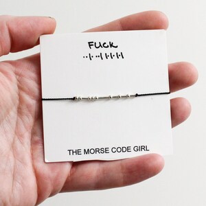 May include: A black string bracelet with silver beads spelling out "FUCK" in Morse code. The bracelet is displayed on a white card with the text "FUCK" and "THE MORSE CODE GIRL".