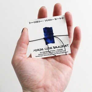May include: A white card with the text "MORSE CODE BRACELET" is held in a hand. The card has a blue watercolor design and is tied with black string. The text reads "An Alphabet of Dots + Dashes" and "Sterling Silver or 14K Gold on Natural Silk Cord."