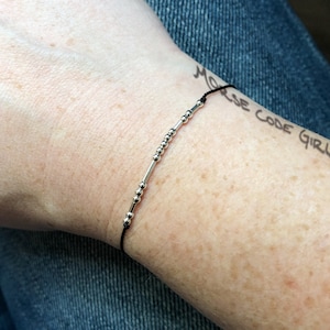 May include: A delicate bracelet with silver beads and cylindrical bars on a black cord. The bracelet is designed with a Morse code pattern. The words "Morse Code Girl" are written on the skin in the background.