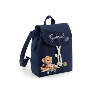 Puede incluir: Una mochila azul marino con un cachorro de león volando un avión. La mochila tiene un cierre de cordón blanco y el nombre "Gabriel" bordado en la parte delantera.