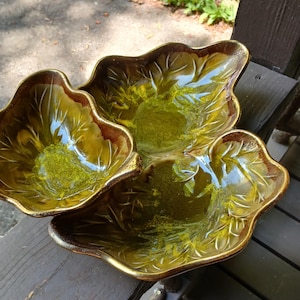 May include: A three-sectioned serving dish in a leaf shape. The dish has a glossy, olive-green interior with brown edges and gold trim. The leaf veins are visible. The dish is likely ceramic or porcelain.