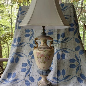 May include: A vintage table lamp with a cream-colored lampshade and a decorative base. The base is a light beige color with floral accents and gold-colored handles. The lamp is set against a blue and white floral patterned backdrop.