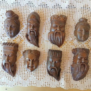 May include: A collection of eight carved wooden wall plaques, each depicting a distinct human face with varied expressions, hairstyles, and headwear. The carvings feature dark brown wood tones with detailed facial features, including beards and hair.