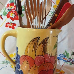 May include: A yellow ceramic pitcher with a fruit design, featuring pears, apples, and bananas. The pitcher is filled with kitchen utensils, including a wooden spoon, a whisk, and a red spatula.