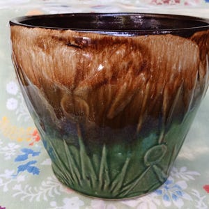 May include: A ceramic planter with a glossy finish. The planter features a gradient of colors, transitioning from dark brown at the rim to a lighter brown and then to green at the base. The base has a relief design of grass and flowers.