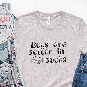 May include: Gray t-shirt with the text "Boys are better in books" and a small illustration of a book. The shirt is displayed on a white surface with a plaid shirt, blue jeans, and brown boots.