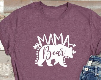 May include: A mauve t-shirt with a white graphic design of a bear with the text "Mama Bear" and arrows. The shirt is laying on a wooden surface with a pair of blue jeans.