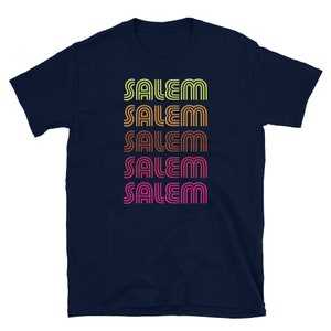 May include: Navy blue t-shirt with the word "SALEM" printed in a repeating pattern. The text is in a retro font, with each instance of the word in a different color: yellow, orange, brown, and pink.