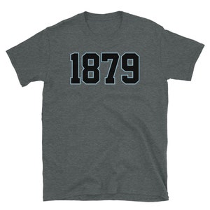 K&ouml;nnte beinhalten: Ein dunkelgraues T-Shirt mit dem Aufdruck 1879 in Schwarz mit hellblauem Umriss.