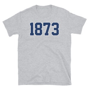 Puede incluir: Una camiseta de manga corta gris jaspeada con el n&uacute;mero 1873 impreso en azul marino.