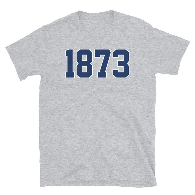 Puede incluir: Una camiseta de manga corta gris jaspeada con el n&uacute;mero 1873 impreso en azul marino.