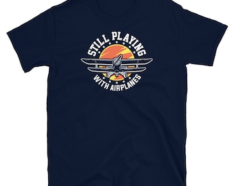 May include: Navy blue t-shirt with a white graphic of a vintage airplane flying through a red and orange sun with the text "Still Playing With Airplanes".