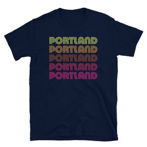May include: A navy blue t-shirt with the word "PORTLAND" repeated five times in a retro, layered font style. Each word is a different color, starting with yellow, then orange, brown, pink, and finally red.