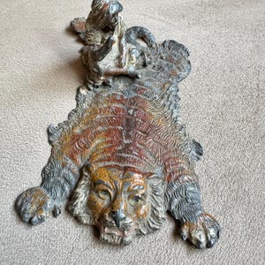 Può includere: Una figurina vintage in metallo di un tappeto di tigre con una scimmia seduta sopra. La tigre è dipinta in tonalità di marrone, nero e arancione, e la scimmia è dipinta in tonalità di grigio e marrone.