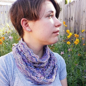 May include: A person with short brown hair wearing a purple and blue knitted cowl neck scarf. The person is standing in a garden with yellow flowers.
