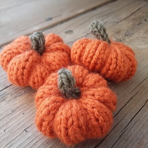Könnte beinhalten: Drei handgefertigte, orangefarbene, gestrickte Kürbisse mit grauen Stielen. Diese dekorativen Kürbisse sind ideal für die Herbstdekoration. Die Kürbisse sind aus weichem, strukturiertem Garn gefertigt.