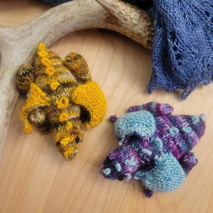 Può includere: Due giocattoli di drago a maglia. Uno è giallo e verde con accenti marroni e l'altro è blu, viola e rosa.