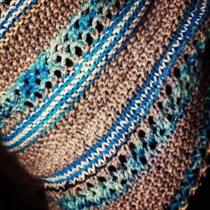 May include: Close-up of a hand-knitted textile with a textured, open weave. The color palette includes shades of gray, blue, and white, with diagonal stripes of blue and white yarn. The knitted fabric has a soft, cozy appearance.