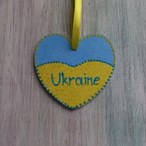 Könnte beinhalten: Ein herzförmiger Holzschmuck, der in den Farben der ukrainischen Flagge, blau und gelb, bemalt ist, mit dem Wort "Ukraine" in Grün auf dem gelben Teil. Der Schmuck hat ein gelbes Band, das oben befestigt ist.