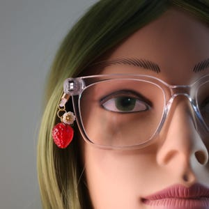May include: Clear-framed eyeglasses with a dangling charm. The charm features a red strawberry bead and small white flower accents. The glasses are worn by a person with green hair.
