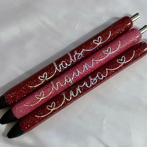 May include: Three red glitter pens with black ink and silver lettering. The pens are labeled "love", "dream", and "believe" with a heart symbol before each word.