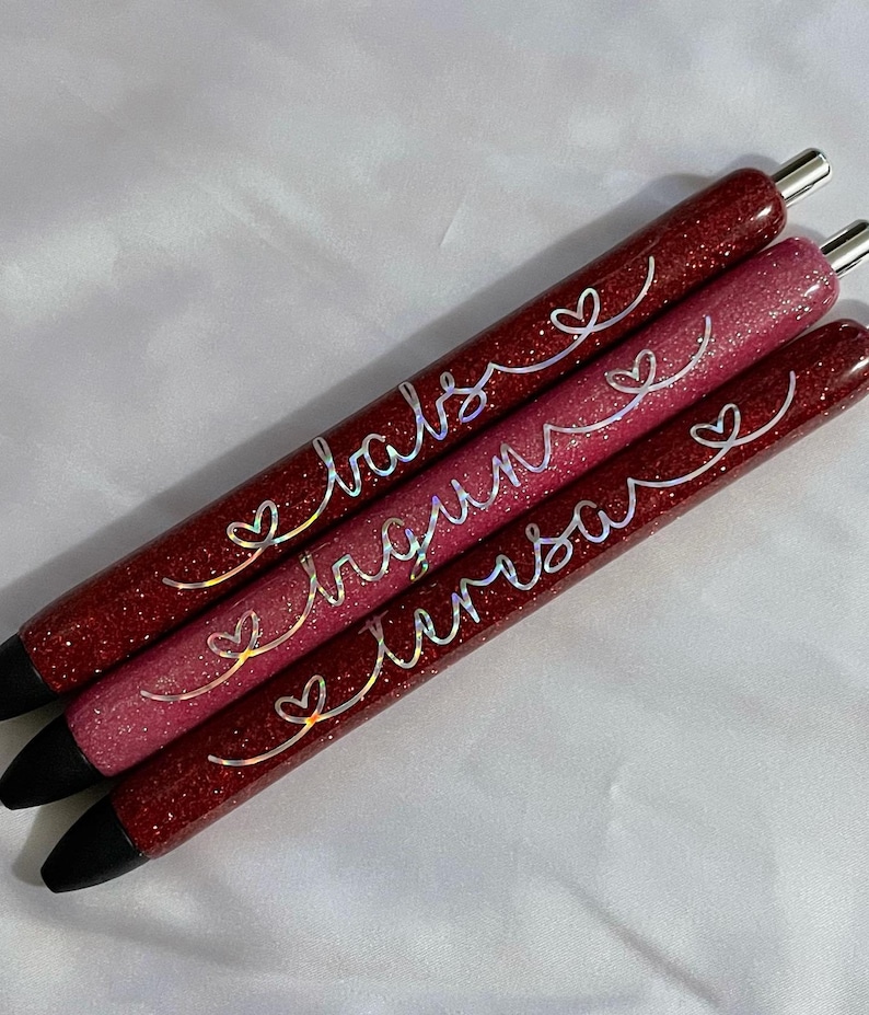 May include: Three red glitter pens with black ink and silver lettering. The pens are labeled "love", "dream", and "believe" with a heart symbol before each word.