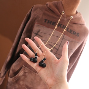 May include: A gold-toned metal chain accessory designed to hold wireless earbuds, featuring delicate star-shaped links. The chain connects to two black wireless earbuds, each with a small white ring detail, providing a secure way to wear audio devices.