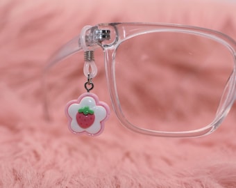 May include: Clear eyeglasses with a pink and white flower charm featuring a strawberry design. The charm is attached to the glasses frame with a small silver-colored chain. The glasses are set against a soft pink background.
