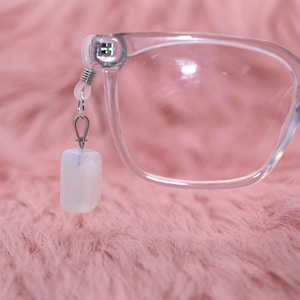 May include: Clear eyeglasses with a rectangular frame and a white, rectangular eyeglass chain charm. The charm is attached to the glasses with a silver-colored chain. The background is a soft, pink, fuzzy surface.