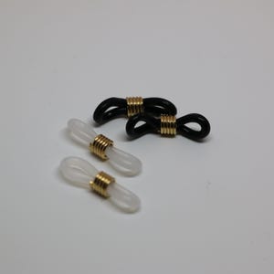 May include: Eyeglass retainers in black and clear with gold-colored metal accents. The retainers are designed to loop around the arms of eyeglasses to keep them secure. The image shows three retainers, two black and one clear.