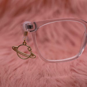 Könnte beinhalten: Ein goldfarbener Planetenanhänger mit Sternakzent hängt an einem transparenten Brillengestell. Der Anhänger ist mit einer kleinen, transparenten Gummischlaufe und goldfarbenen Perlen an der Brille befestigt. Der Hintergrund ist eine weiche, rosa, verschwommene Textur.