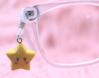 Könnte beinhalten: Eine Brille mit transparentem Rahmen und einem gelben Sternanhänger, der am Rahmen hängt. Der Stern hat ein cartoonartiges Design mit einem lächelnden Gesicht und ist mit einer silberfarbenen Metallschlaufe an der Brille befestigt.