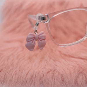 May include: A close-up of eyeglasses with a pink bow-shaped charm attached to the frame. The glasses are clear, and the bow is a soft, pastel pink. The background is a fuzzy, pink surface.