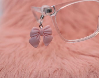 May include: A close-up of eyeglasses with a pink bow-shaped charm attached to the frame. The glasses are clear, and the bow is a soft, pastel pink. The background is a fuzzy, pink surface.