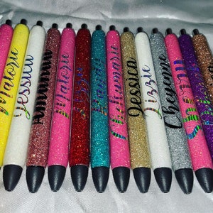 May include: A set of 12 glitter pens with personalized names. The pens are in various colors, including yellow, white, pink, red, blue, gold, silver, and purple. Each pen has a black ink tip.