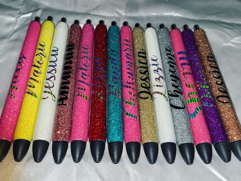 May include: A set of 12 glitter pens with personalized names. The pens are in various colors, including yellow, white, pink, red, blue, gold, silver, and purple. Each pen has a black ink tip.