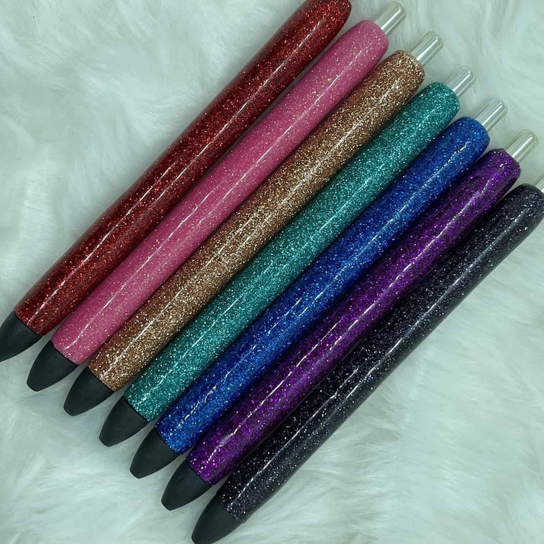 May include: Seven glitter pens in various colors, including red, pink, gold, teal, blue, purple, and black. The pens have clear tops and are arranged in a row.
