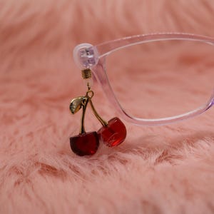 Könnte beinhalten: Eine Brille mit einem hellvioletten Rahmen und einem Kirschanhänger. Der Anhänger zeigt zwei rote Kirschen mit goldenen Stielen und einem goldenen Blatt. Der Hintergrund ist eine weiche, rosa, pelzige Textur.
