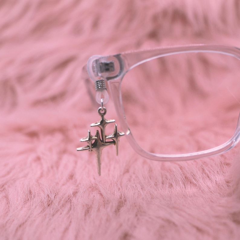 May include: Clear eyeglasses with a silver starburst charm hanging from the frame. The charm features multiple star shapes of varying sizes. The background is a soft, pink, fuzzy texture.