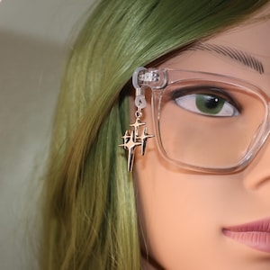 May include: A gold-colored eyeglass chain charm featuring a cluster of star-shaped pendants. The charm is attached to a pair of clear-framed eyeglasses. The background includes green hair and a mannequin face.