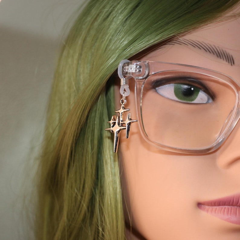May include: A gold-colored eyeglass chain charm featuring a cluster of star-shaped pendants. The charm is attached to a pair of clear-framed eyeglasses. The background includes green hair and a mannequin face.