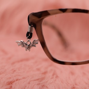 Peut inclure: Un charm chauve-souris argenté, suspendu à un cordon noir, attaché à la branche d'une paire de lunettes. Les lunettes présentent un motif écaille de tortue marron et beige. Le fond est une surface rose douce et texturée.