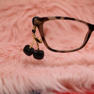 May include: Eyeglasses with a tortoiseshell pattern frame. A gold-coloured charm with two black cherries hangs from the frame. The background is a soft, pink, fuzzy texture.