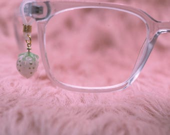 May include: Clear eyeglasses with a small strawberry charm attached to the frame. The strawberry charm is white with green accents and gold details. The glasses are set against a soft, pink background.