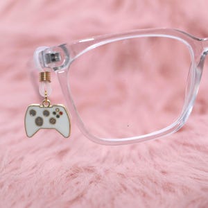 May include: Clear-framed eyeglasses with a white and gold-colored video game controller charm hanging from the frame. The controller has gray buttons and red and green accents. The background is a soft, pink, fuzzy material.