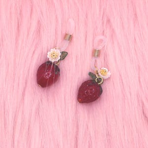 May include: Two pairs of eyeglass chains with strawberry charms. The charms are red with green leaves and small white flowers. The chains are clear and have gold-colored metal accents. The background is pink.