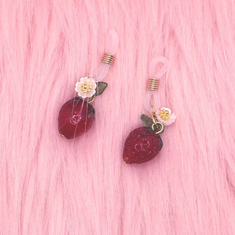 May include: Two pairs of eyeglass chains with strawberry charms. The charms are red with green leaves and small white flowers. The chains are clear and have gold-colored metal accents. The background is pink.